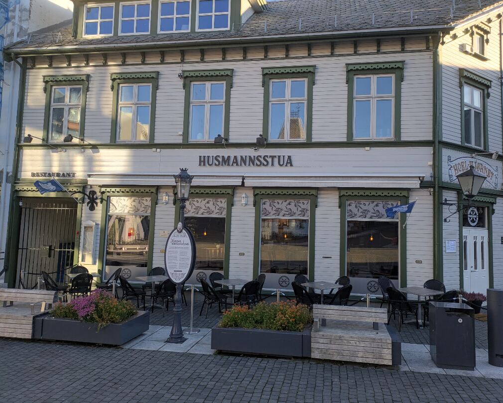 Fasaden med skiltet «Husmannsstua», uteservering med svarte stoler og blomsterkasser foran vinduene